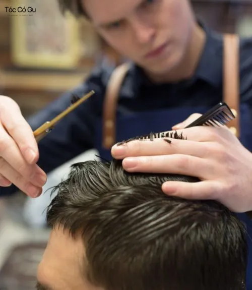 Tóc Side Part là gì? Top 10 kiểu tóc Side Part quốc dân hiện nay Thợ cắt tóc đang dùng lược và kéo tỉa phần mái cho nam giới tại tiệm barber.